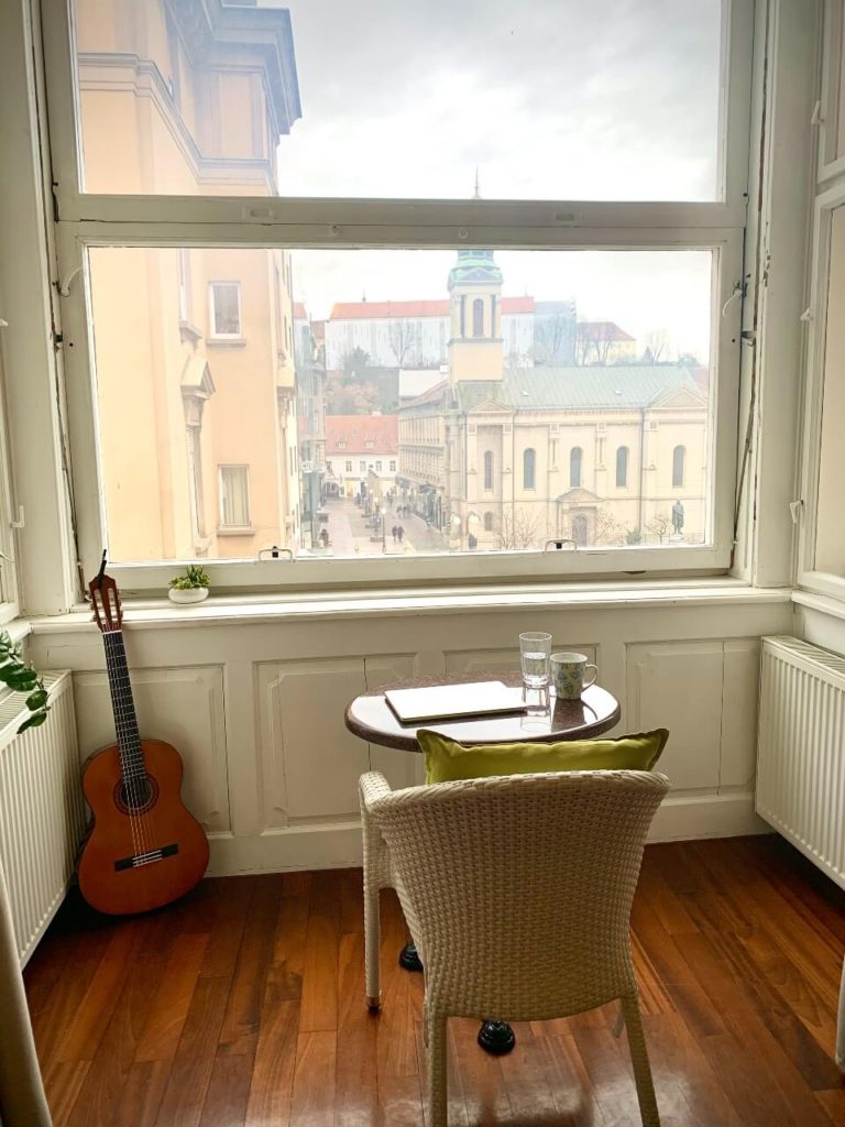 A view from Leah's apartment overlooking the square in Zagreb. The floors are hardwood, the walls around the windows are white, there's a table in the center with a coffee cup and a guitar resting in the corner.